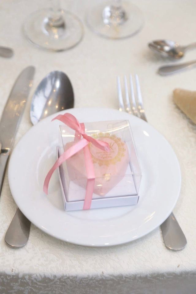 Pink heart-shaped wedding favour cake with a gold emblem in a clear ribboned gift box, set on a place setting