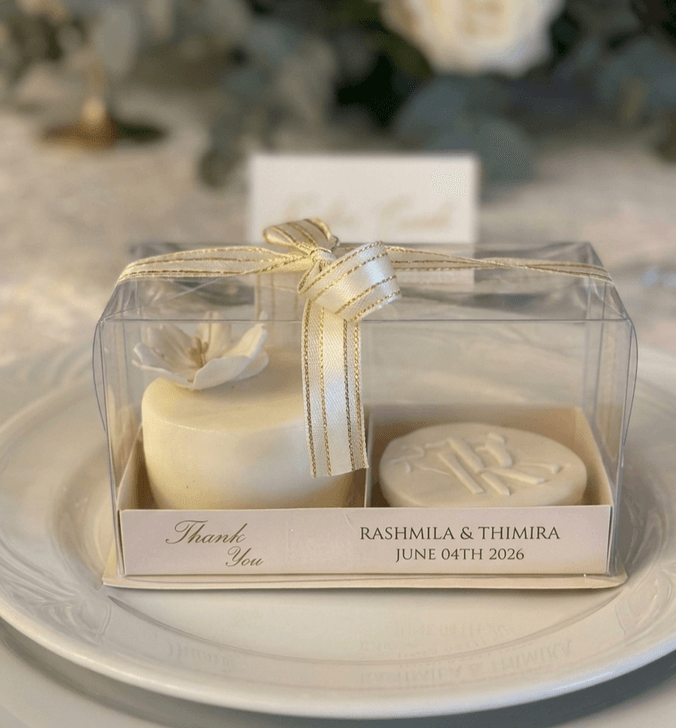 Ivory wedding favour set — a white chocolate mini cake with a sugar flower and a name-embossed butter cookie — in a clear custom gift box tied with a gold-edged ribbon, by Cake by O2 in Colombo