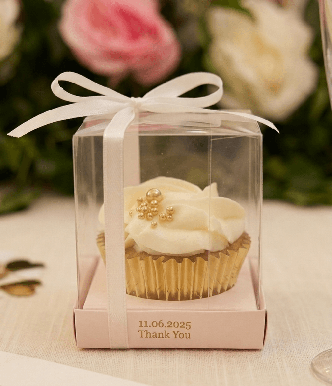 Vanilla wedding favour cupcake with swirled buttercream and gold pearls in a clear thank-you gift box, with roses behind