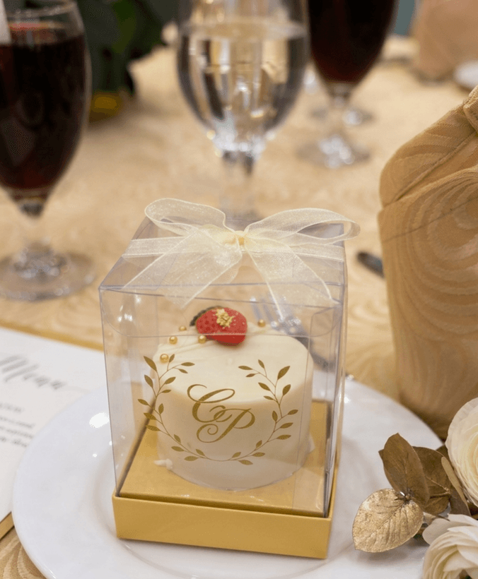White wedding favour mini cake with a gold monogram and leaf wreath, topped with a strawberry, in a clear gift box
