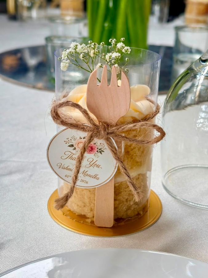 Naked vanilla mini cake with exposed sponge and a buttercream swirl in a clear cup on a gold base, with a wooden fork, twine bow, baby's breath and a personalised thank-you tag, by Cake by O2 in Colombo