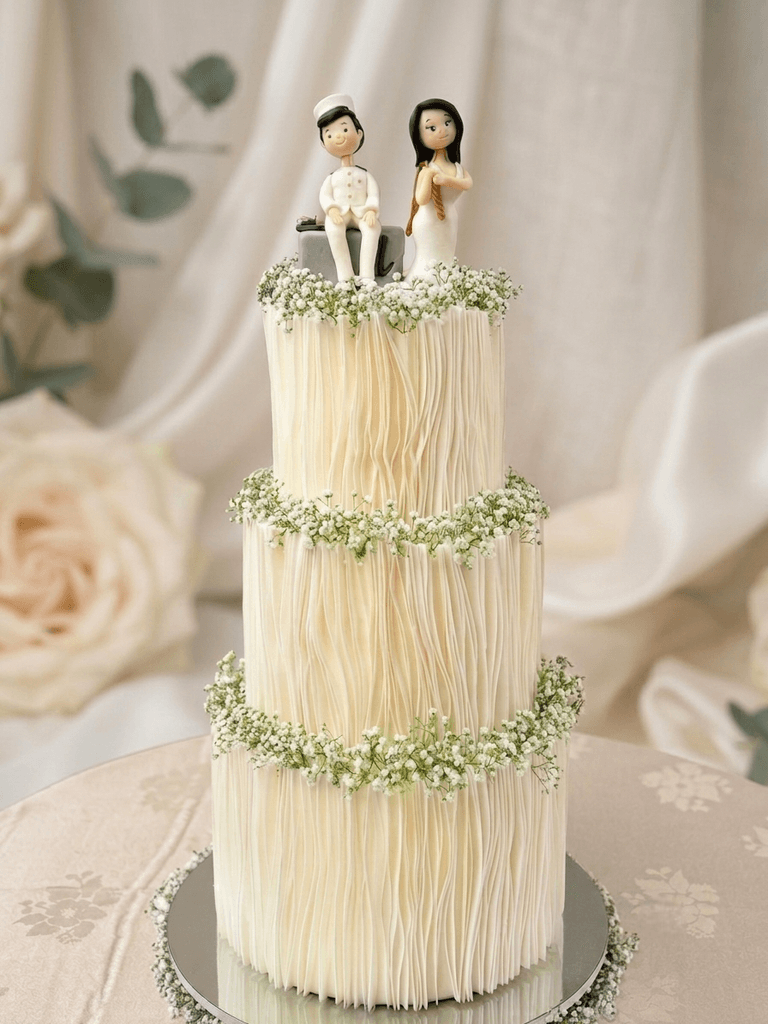Three-tier white wedding cake with wafer-paper pleats, baby's breath garlands and a hand-modelled couple topper by Cake by O2, Colombo