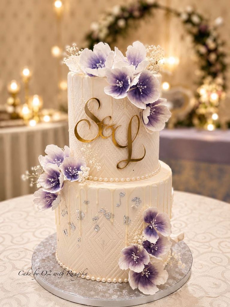 Two-tier ivory wedding cake with purple ombré sugar flowers, a gold S&L monogram and pearl trim, by Cake by O2 in Colombo