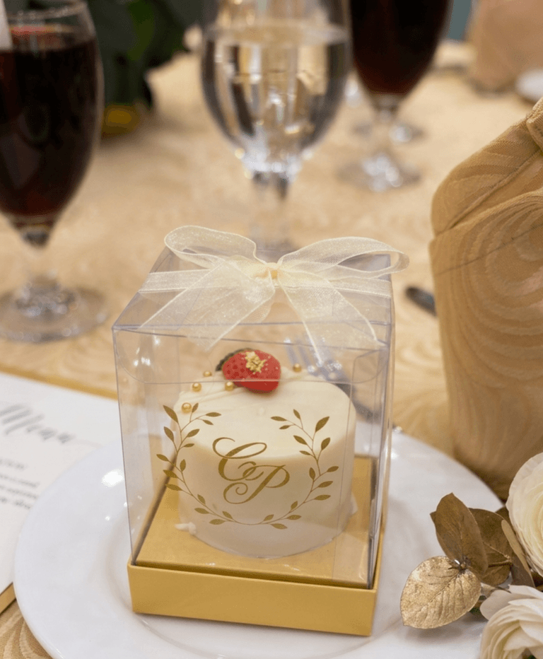 White wedding favour mini cake with a gold monogram and leaf wreath, topped with a strawberry, in a clear gift box